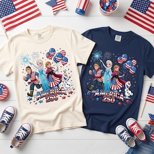 K&ouml;nnte beinhalten: Zwei kurz&auml;rmelige T-Shirts, eines in Creme und eines in Marineblau, mit einem Grafikdruck von drei animierten Figuren und einem Schneemann. Das Design zeigt US-Flaggenmuster, Luftballons, Feuerwerk und den Schriftzug "AMERICA 250" in Rot, Wei&szlig; und Blau.
