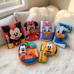 May include: Six letter-shaped plush pillows featuring Disney characters. The letter M shows Mickey Mouse and the name Matthew. The letter C features Minnie Mouse and Charlotte. D shows Daisy Duck and Daniel. E shows Donald Duck and Emily. G shows Goofy and Grace. P shows Pluto and Patrick.