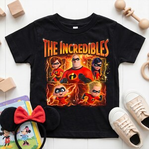 May include: A black short-sleeved t-shirt featuring a graphic print of the characters from The Incredibles. The design includes the title text in orange and yellow, with the family members in their red suits against a background of orange lightning.