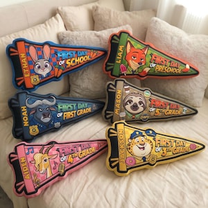 Puede incluir: Una colecci&oacute;n de almohadas en forma de bander&iacute;n con personajes de Zootopia. Cada almohada tiene un nombre y un grado escolar, como "First Day of School" y "First Grade". Las almohadas est&aacute;n sobre una cama blanca.