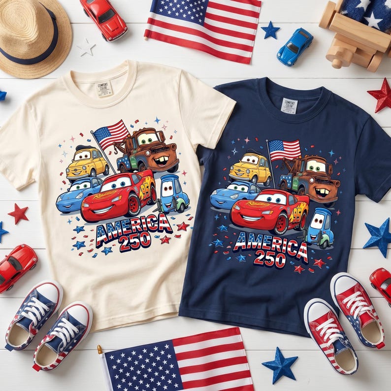 May include: A cream and a navy blue t-shirt, each featuring a graphic print of animated cars including a red race car, a brown tow truck, and a yellow car, with the text AMERICA 250, surrounded by small stars and a United States flag.