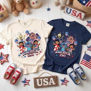 May include: Two short-sleeve t-shirts, one cream and one navy blue, each featuring a graphic of Lilo, Stitch, and other characters holding American flag balloons. The text "AMERICA 250" is printed below the characters. The shirts are surrounded by small American flag stars and two wooden signs labeled "USA".