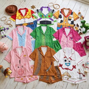 May include: A collection of short-sleeved button-down pajama sets featuring designs inspired by various toy characters. The sets include patterns resembling a space ranger, a cowboy, a cowgirl, a pink bear, a green dinosaur, a pig, a dog, and a spork.