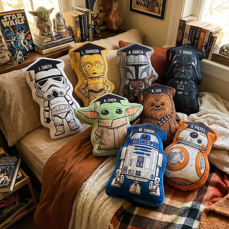 May include: A collection of Star Wars character-themed pillows, including Stormtrooper, C-3PO, The Mandalorian, Darth Vader, Chewbacca, Grogu, R2-D2, and BB-8. Each pillow is shaped like the character and wears a graduation cap with a name.