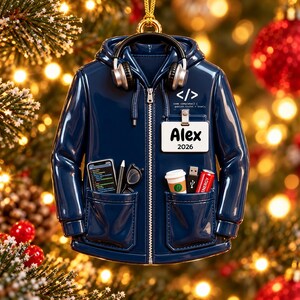 Puede incluir: Un adorno de sudadera con capucha azul marino con cremallera, auriculares negros, una tarjeta de identificaci&oacute;n blanca con el nombre Alex 2026 y c&oacute;digo. Los bolsillos contienen un smartphone, bol&iacute;grafo, gafas, caf&eacute;, USB y una lata roja.