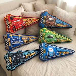 May include: A collection of colorful, car-shaped pillows. Each pillow features a different car character and the text "First Day of School" or a grade level. The pillows are in various colors, including red, blue, green, and brown.