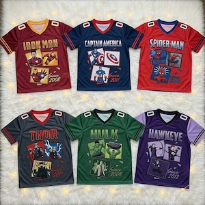 May include: Six V-neck jerseys featuring Marvel characters. Top row: red Iron Man, blue Captain America, red Spider-Man. Bottom row: grey Thor, green Hulk, purple Hawkeye. Each shirt displays character graphics, names, and debut years.