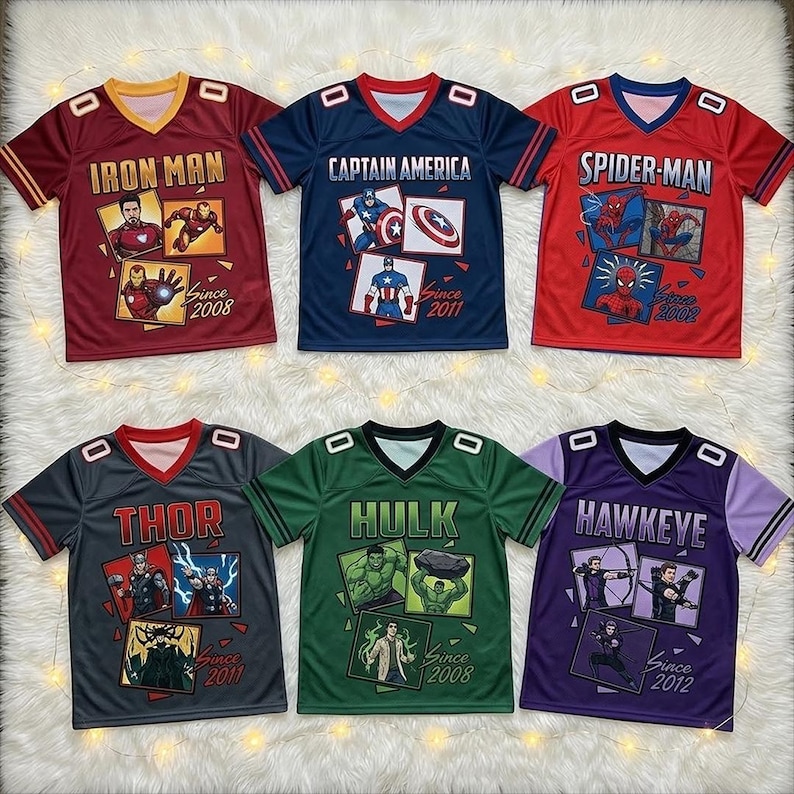 May include: Six V-neck jerseys featuring Marvel characters. Top row: red Iron Man, blue Captain America, red Spider-Man. Bottom row: grey Thor, green Hulk, purple Hawkeye. Each shirt displays character graphics, names, and debut years.