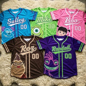 May include: Baseball jerseys in blue, green, pink, brown, and purple, each featuring a character from the animated film. The jerseys have character names and the number "00" on the front. The jerseys are displayed on a fluffy, light-colored surface.