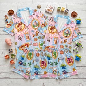 May include: Two sets of short-sleeved button-down pajamas featuring a light blue and pink gradient background with a repeating Toy Story character print. The pattern includes Woody, Buzz Lightyear, and Lotso in gold frames, along with alphabet blocks.
