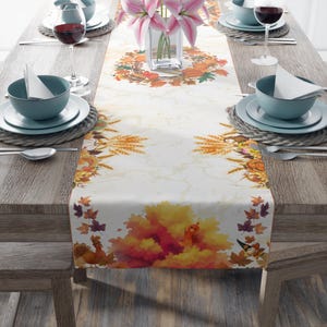 Pode incluir: Um caminho de mesa branco com uma estampa festiva de outono com abóboras, talos de trigo, folhas coloridas e perus. O caminho está sobre uma mesa de madeira posta com tigelas azuis, guardanapos brancos, taças e um vaso de vidro com lírios.