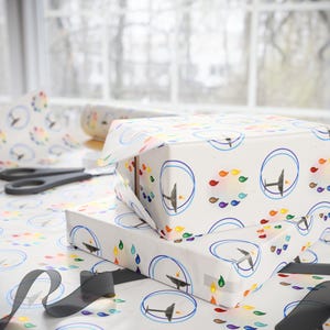 May include: Two gift boxes wrapped in white paper with a repeating pattern of blue circles, colorful teardrops, and a stylized airplane. A roll of matching wrapping paper, scissors, and black ribbon are also visible.