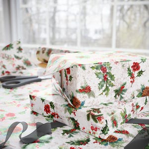 May include: A rectangular gift box wrapped in white paper featuring a watercolor print of green holly leaves, red berries, and brown pinecones. A pair of black scissors and a black ribbon are placed on the patterned wrapping paper surface.