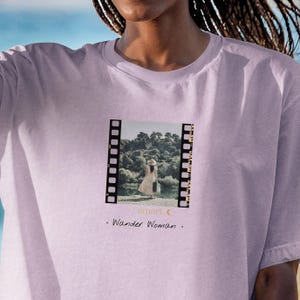 Könnte beinhalten: Helllilafarbenes T-Shirt mit einem Filmstreifen-Motiv. Die Grafik zeigt eine Frau in einem Kleid und Hut am Wasser, mit dem Text "amart" und "Wander Woman".