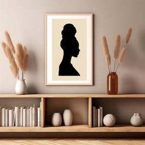 May include: A black and white silhouette of a woman with her hair in a bun, printed on a beige background. The print is framed in a light wood frame and is displayed on a shelf with books and vases.