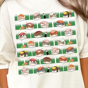 May include: Ivory-colored t-shirt featuring a grid of illustrated sushi rolls on a green and white background. The sushi designs include various types, with details like rice, seaweed, and toppings. The word "IVORY" is printed at the bottom left.