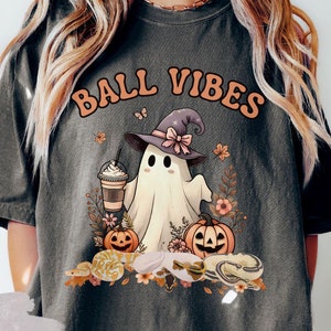 May include: Dark grey t-shirt with a Halloween-themed graphic. The design features a cartoon ghost wearing a witch hat, holding a coffee, and surrounded by pumpkins and flowers. The text "BALL VIBES" is arched above the graphic.