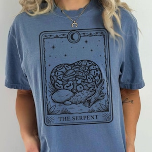 May include: Blue jean t-shirt featuring a black graphic of a snake within a tarot card design. The card reads "THE SERPENT" at the bottom. The shirt is a relaxed fit.