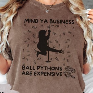 May include: Espresso-colored t-shirt with the text "MIND YA BUSINESS" and "BALL PYTHONS ARE EXPENSIVE." The graphic features a silhouette of a person pole dancing, surrounded by dollar bills. A snake graphic is also present.
