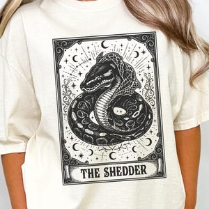 May include: Ivory-colored t-shirt featuring a black and white illustration of a snake within a decorative frame. The design includes celestial elements and the text "THE SHEDDER" at the bottom. The shirt is a relaxed fit.