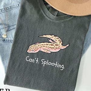 Può includere: Una maglietta grigio scuro con un'illustrazione di un geco dei cartoni animati e la scritta "Can't. Splooting." La maglietta è appoggiata su jeans blu e un cappello beige.