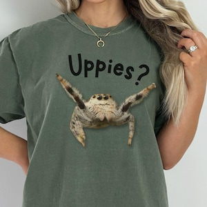 Può includere: T-shirt verde muschio con la scritta "Uppies?" sopra un'illustrazione dettagliata di un ragno saltatore. Il ragno è marrone e beige con le zampe divaricate. La parola "moss" è in basso.