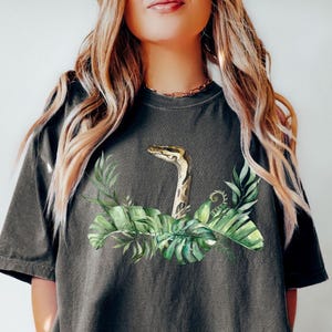 May include: Dark gray t-shirt featuring a watercolor illustration of a snake with a green and yellow pattern, surrounded by green tropical leaves. The word "PEPPER" is printed in black at the bottom left.