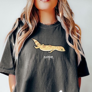 Può includere: T-shirt grigio scuro con un geco dei cartoni animati e la parola "nope". Il geco è marrone chiaro con macchie marroni. La parola "pepper" è stampata in basso a sinistra nell'immagine.