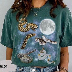 May include: A teal blue t-shirt with a galaxy print featuring five snakes in different poses. The snakes are a mix of brown, yellow, and white. The shirt has a round neckline and short sleeves.