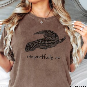 Può includere: T-shirt color espresso con un'illustrazione dettagliata di un geco e la frase "respectfully, no." Il design del geco è nei toni del marrone e del nero. È un capo d'abbigliamento casual.