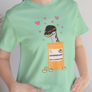 May include: A mint green t-shirt with a cartoon snake in a pill bottle that says "Antidepressant" and "Take as needed daily". The snake is surrounded by pink hearts and gold stars.