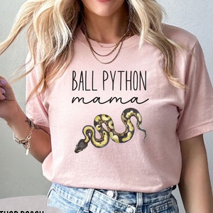 May include: A pink t-shirt with the text "BALL PYTHON mama" and a graphic of a ball python snake. The shirt is made of a soft, comfortable material and is available in Heather Peach.