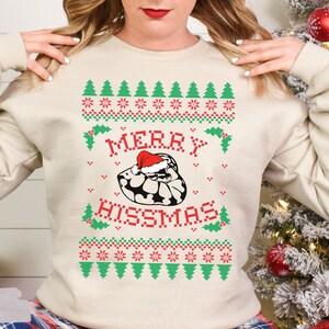 May include: A cream-colored sweatshirt with a festive Christmas design featuring a snake wearing a Santa hat and the text "Merry Hisssmas".