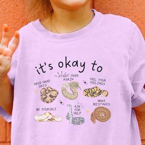 May include: A light purple t-shirt with the text "it's okay to" and various illustrations. The shirt also has the word "ORCHID" printed at the bottom. The shirt is being worn by a young person.