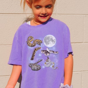 May include: A purple t-shirt with a graphic of snakes and a moon. The snakes are in various poses and are a light brown color. The moon is white and has a textured surface. The t-shirt is a relaxed fit and has a crew neck.