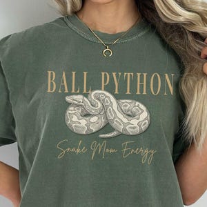 May include: Moss green t-shirt with the text "BALL PYTHON" above a graphic of a snake. Below the snake graphic is the text "Snake Mom Energy". The shirt is a comfortable, casual style.