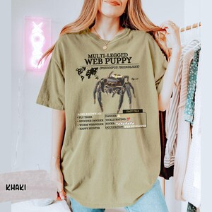 May include: A khaki t-shirt with a graphic of a spider and the text "MULTI-LEGGED WEB PUPPY (PHIDDIPUS FRIENDLIEST)". The text also includes "ALBO KNOWN AS: FLY TIGER, SPOODER DOODER, WORM WRANGLER, HAPPY HUNTER" and "DANGER: TICKLE RATING: SOCKS, OCCUPATION: WEB DEVELOPER".