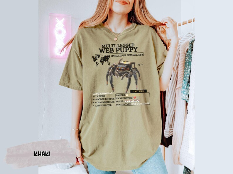 May include: A khaki t-shirt with a graphic of a spider and the text "MULTI-LEGGED WEB PUPPY (PHIDDIPUS FRIENDLIEST)". The text also includes "ALBO KNOWN AS: FLY TIGER, SPOODER DOODER, WORM WRANGLER, HAPPY HUNTER" and "DANGER: TICKLE RATING: SOCKS, OCCUPATION: WEB DEVELOPER".