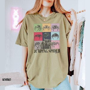 Op de afbeelding: Een kaki t-shirt met een afbeelding van springende spinnen in kleurrijke vierkanten. De tekst "I'm a Jumping Spider" is onder de afbeelding gedrukt.