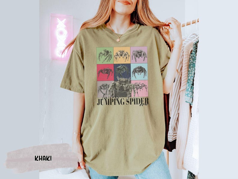 Op de afbeelding: Een kaki t-shirt met een afbeelding van springende spinnen in kleurrijke vierkanten. De tekst "I'm a Jumping Spider" is onder de afbeelding gedrukt.