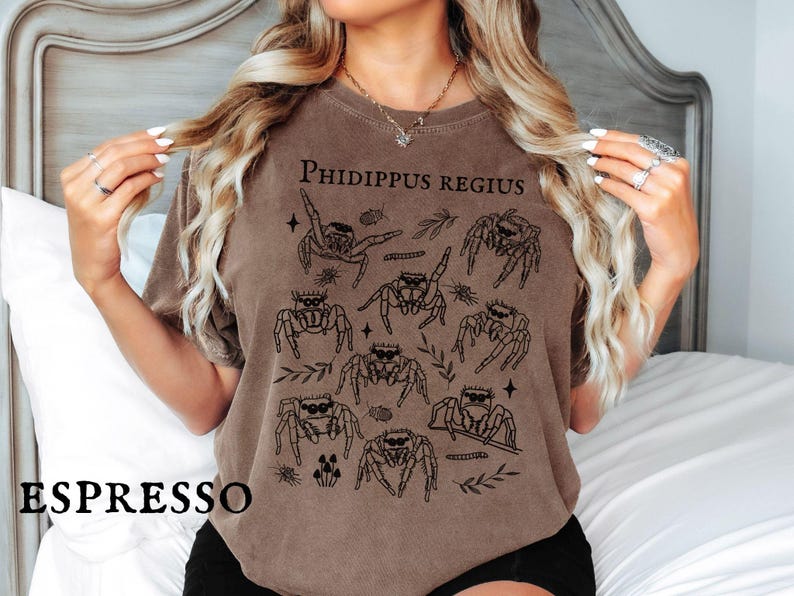 May include: Espresso-colored t-shirt featuring a graphic of several jumping spiders with the text "PHIDIPPUS REGIUS". The design includes botanical elements and stars. The shirt is a comfortable, casual style.