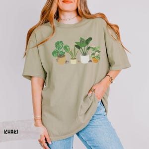 May include: A khaki green t-shirt with a graphic print of five potted plants. The plants are green and have brown pots. The t-shirt is oversized and has a relaxed fit.