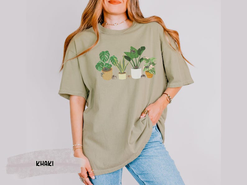 May include: A khaki green t-shirt with a graphic print of five potted plants. The plants are green and have brown pots. The t-shirt is oversized and has a relaxed fit.