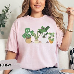 May include: A pink t-shirt with a graphic of five potted plants and spiders. The plants are a monstera, a snake plant, a dracaena, a peace lily, and a kalanchoe. The text "BLOSSOM" is visible in the lower left corner of the image.