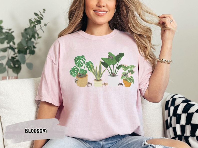 May include: A pink t-shirt with a graphic of five potted plants and spiders. The plants are a monstera, a snake plant, a dracaena, a peace lily, and a kalanchoe. The text "BLOSSOM" is visible in the lower left corner of the image.
