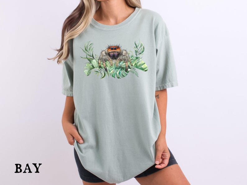 Puede incluir: Una camiseta verde claro con una ilustraci&oacute;n de acuarela de una ara&ntilde;a saltarina rodeada de hojas verdes. La palabra "BAY" est&aacute; impresa en negro en la parte inferior izquierda de la camiseta.