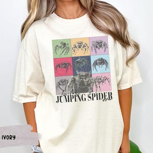 Op de afbeelding: Een wit T-shirt met een kleurrijke afbeelding van springende spinnen en de tekst "I'm a Jumping Spider girl".