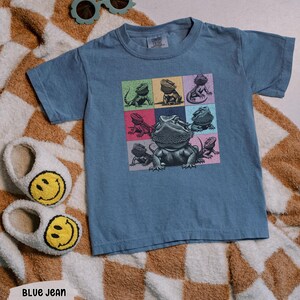 Pu&ograve; includere: Una t-shirt blu jeans con una grafica di diversi draghi barbuti in quadrati colorati. La t-shirt ha un collo rotondo e maniche corte.