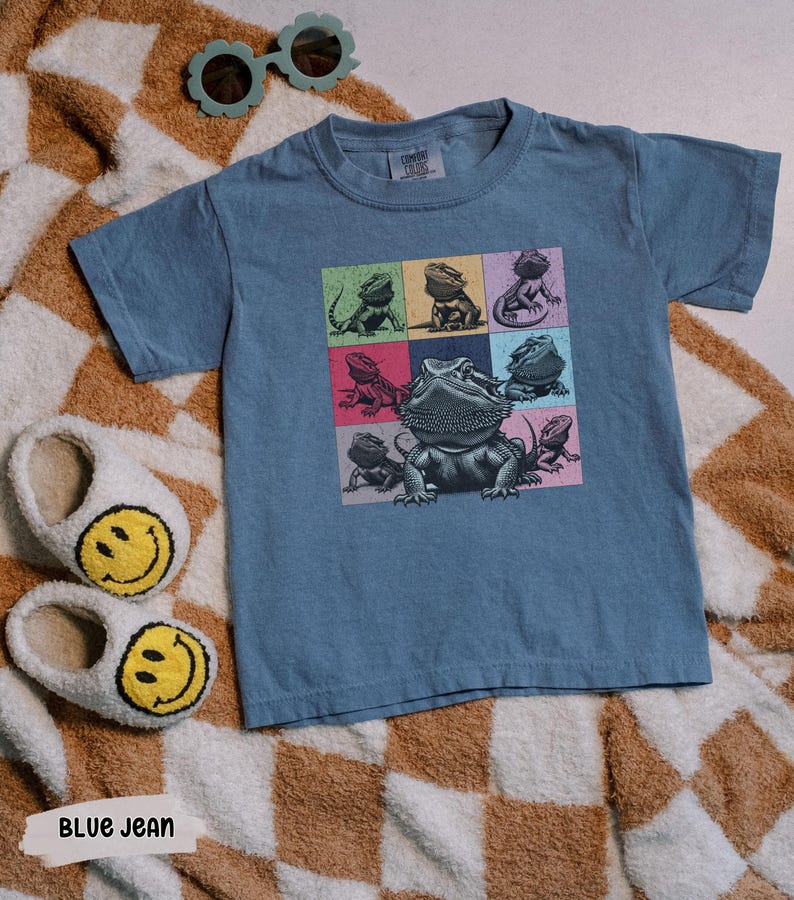 Pu&ograve; includere: Una t-shirt blu jeans con una grafica di diversi draghi barbuti in quadrati colorati. La t-shirt ha un collo rotondo e maniche corte.
