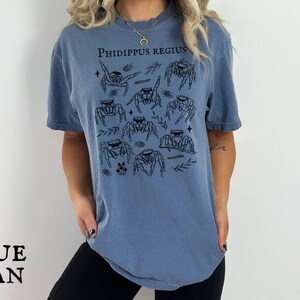 Pu&ograve; includere: T-shirt blu jeans con illustrazione nera di ragni saltatori e la scritta "PHIDIPPUS REGIUS". La maglietta ha una vestibilit&agrave; ampia e maniche corte, adatta per un abbigliamento casual.